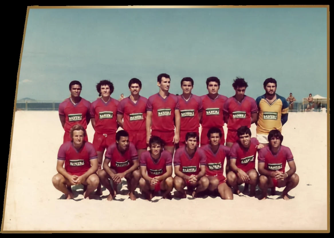 Grupo de jogadores com uniformes vermelhos, posando em duas fileiras na areia.
